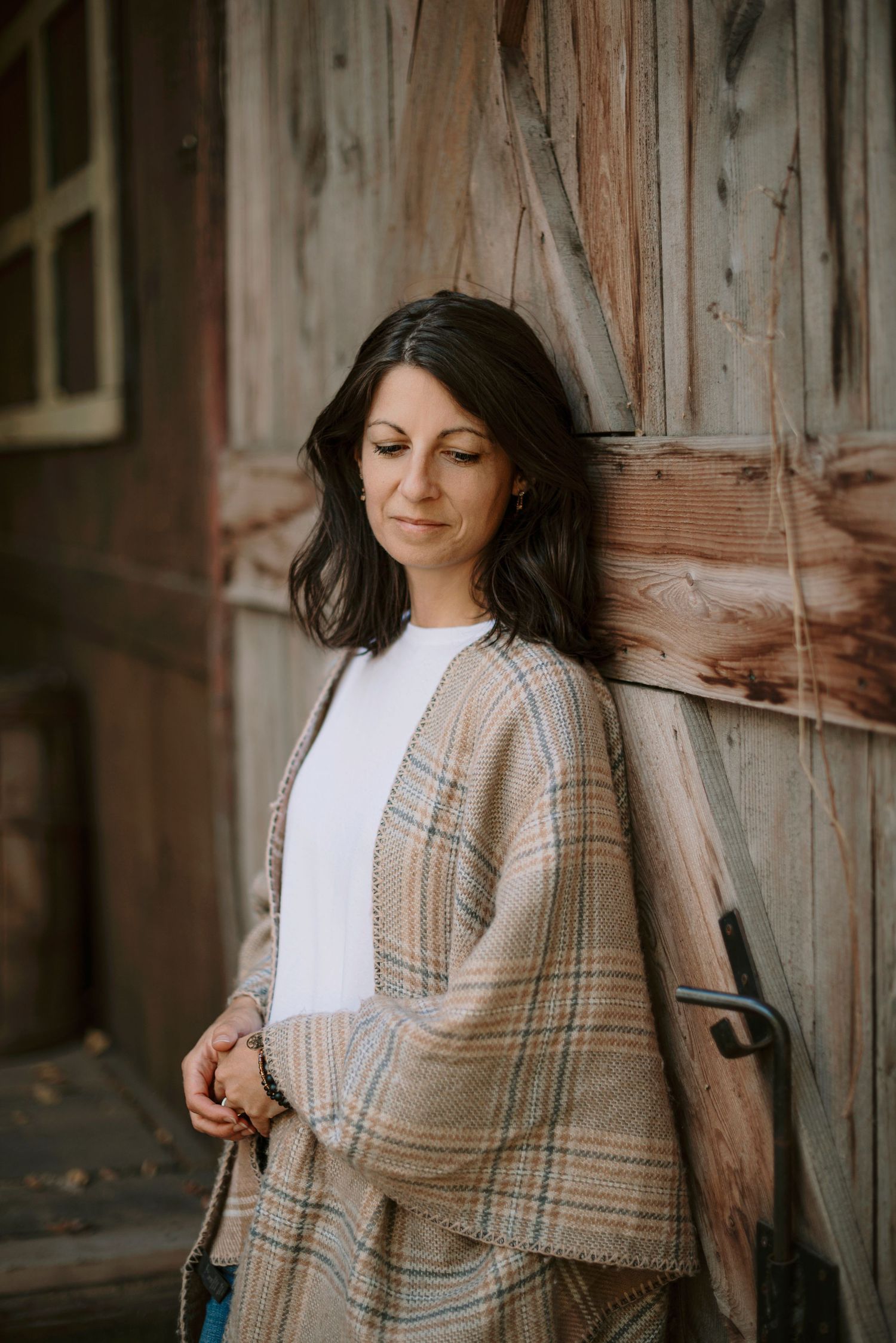 A person in a beige plaid cardigan and white top leans against a rustic wooden wall while smiling warmly.