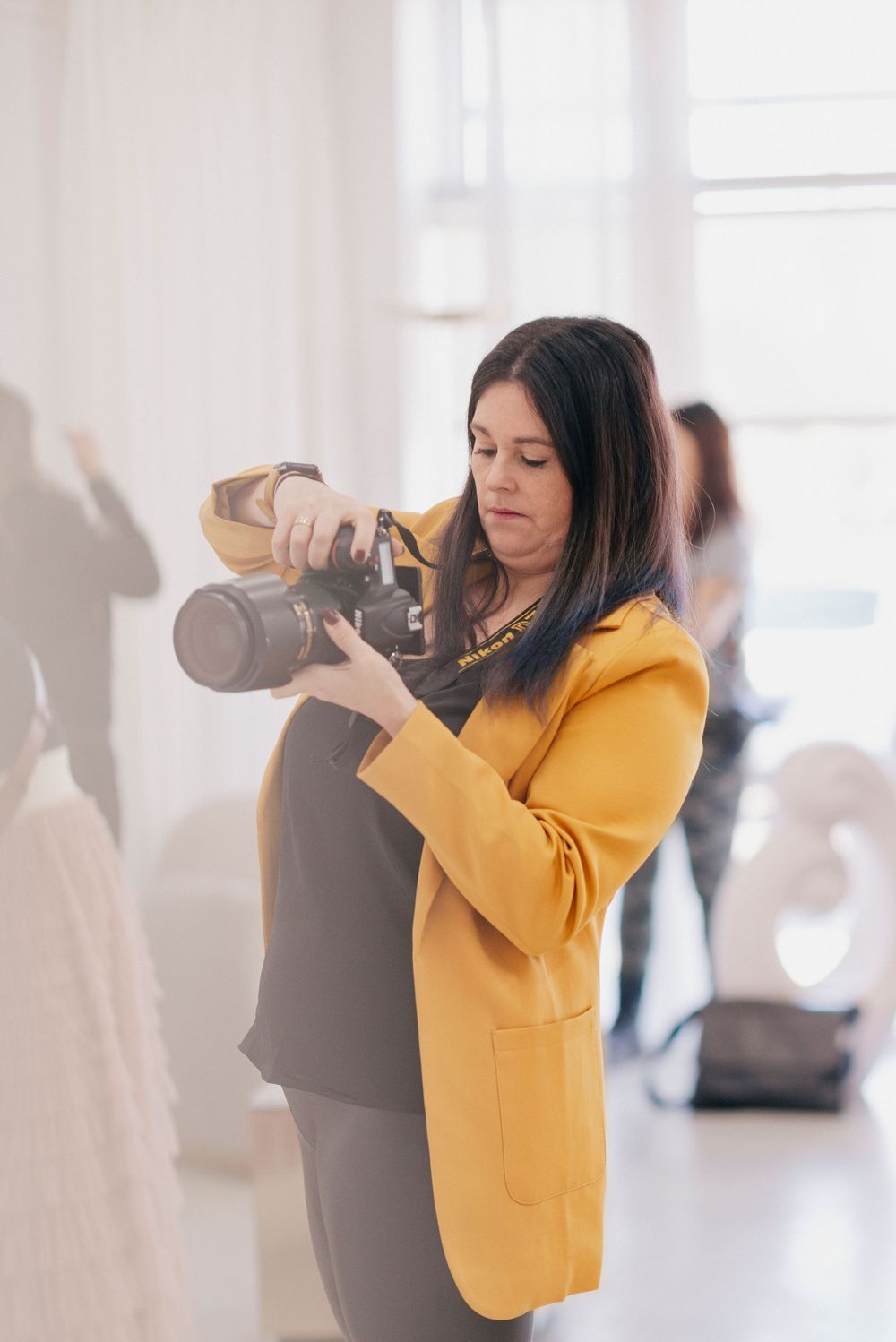 Professional photographer wearing yellow blazer takes photos with DSLR camera in bright indoor studio space.