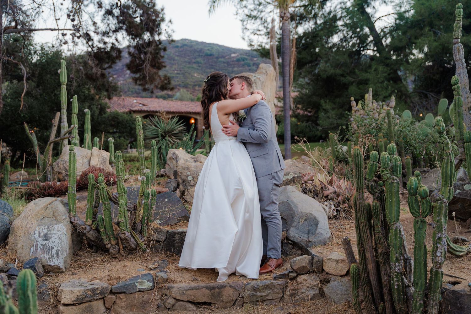 Couple sharing first kiss surrounded by desert cacti and rocks at outdoor wedding venue.