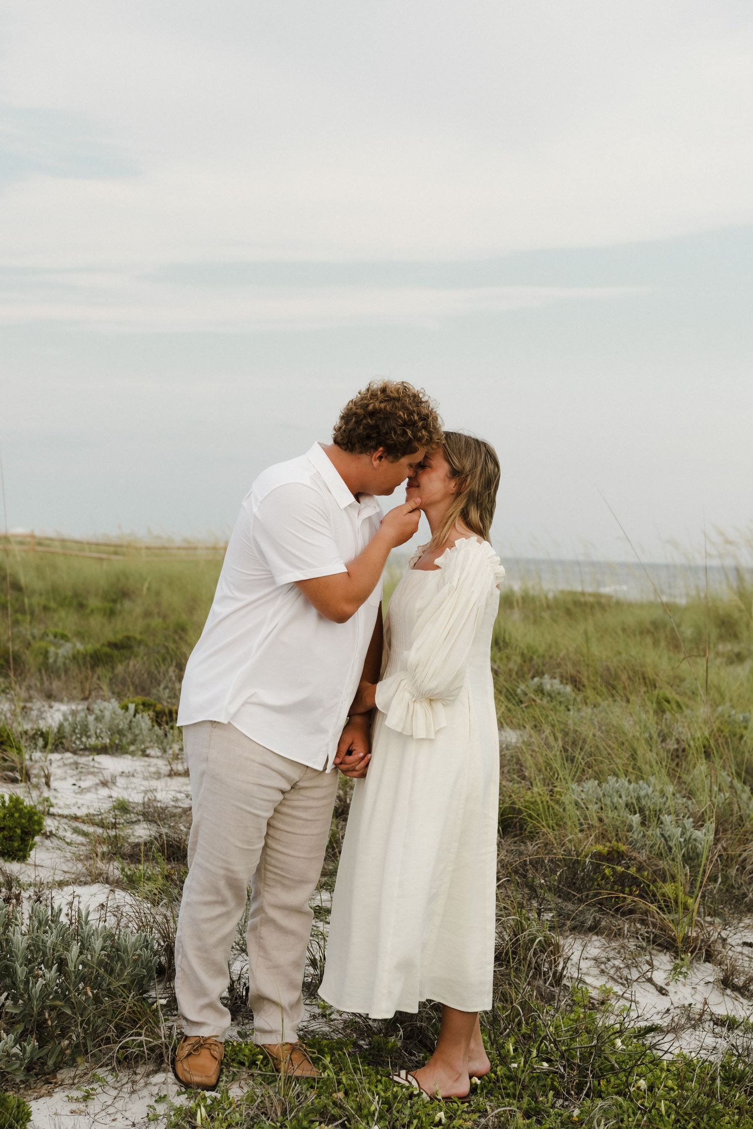 Young couple in white clothing shares tender moment on coastal dunes.