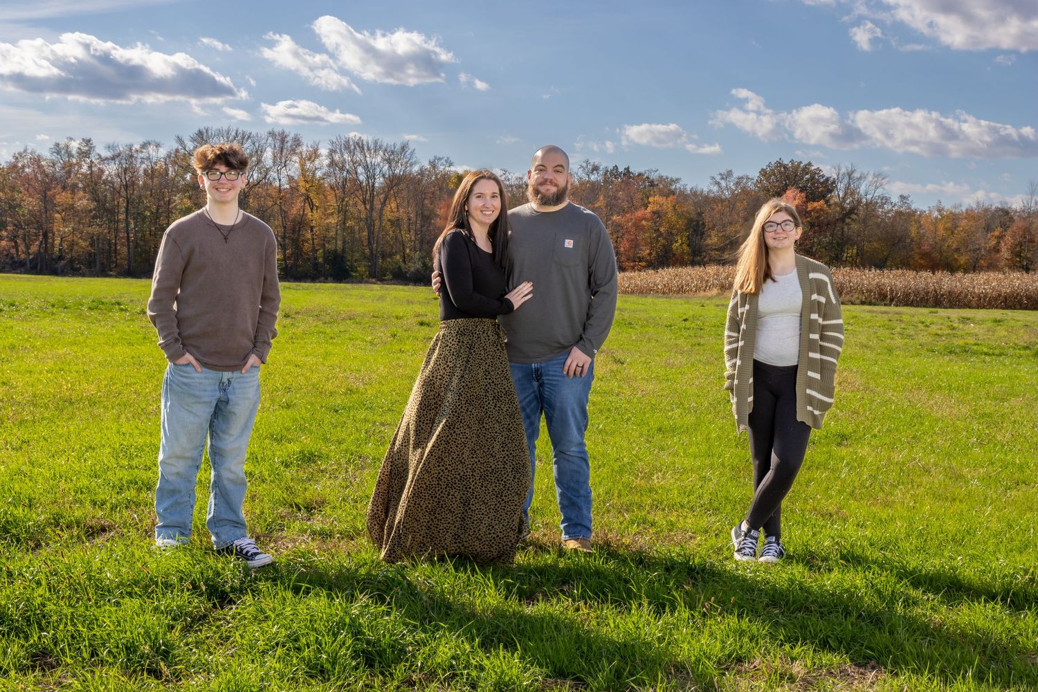 Family of four posing in a green field with autumn trees and blue sky in the background. Salem and Gloucester County Family Portraits