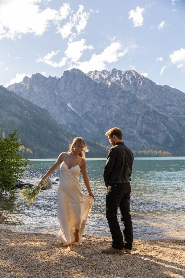 Bride and groom stand by a mountain lake shore, she holds wildflowers in a white dress, majestic peaks behind them.