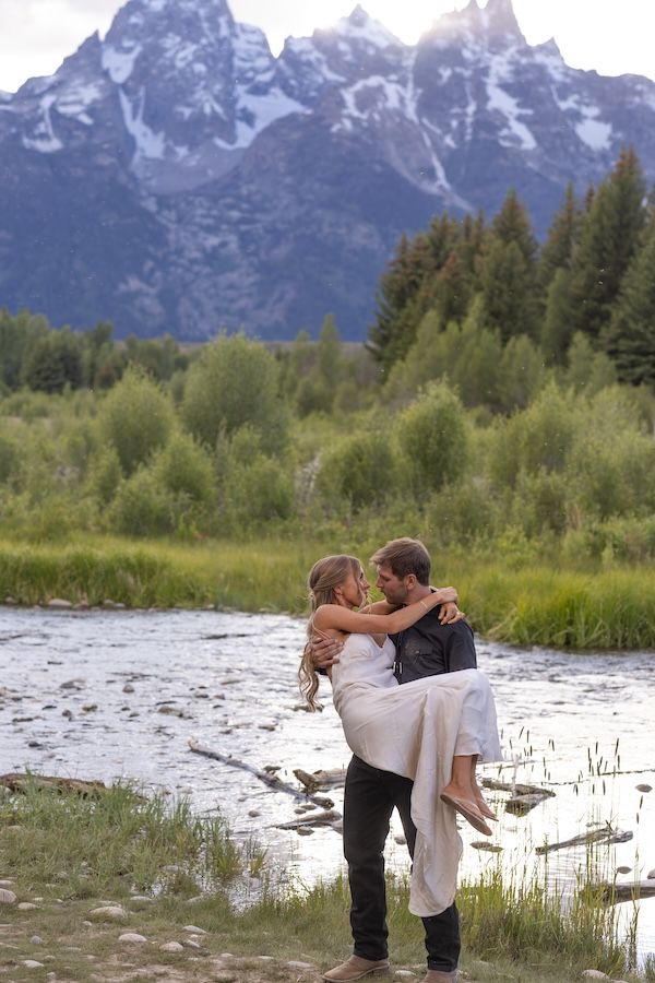 Groom carries bride in white dress by a mountain river with Grand Teton peaks and green trees in background.