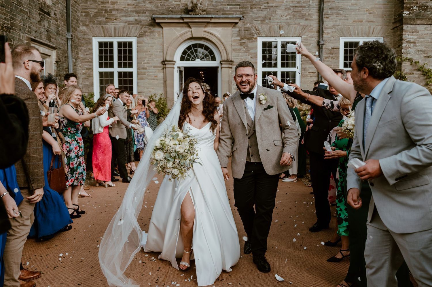 A wedding couple poses together on a gravel path in front of a grand stone archway entrance at sunset at Badminton Court.