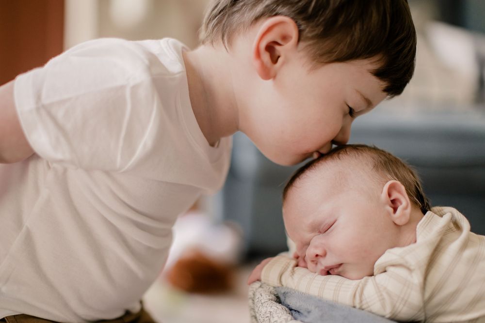 Young child leans in to give a gentle kiss to a sleeping baby wrapped in white swaddle blanket.