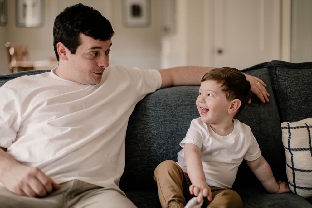 A young boy in a white t-shirt sits on a gray couch and makes playful expressions in a sequence of photos.