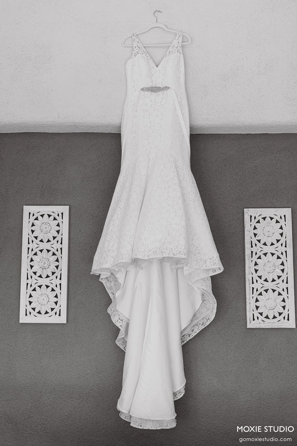 A white wedding dress with lace details hangs against a gray wall with decorative white air vents.