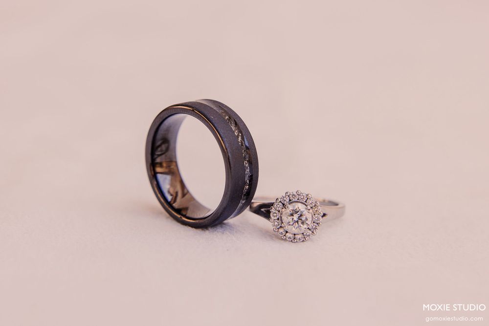 Two wedding rings displayed on a light pink background, one black band and one silver ring with a diamond setting.
