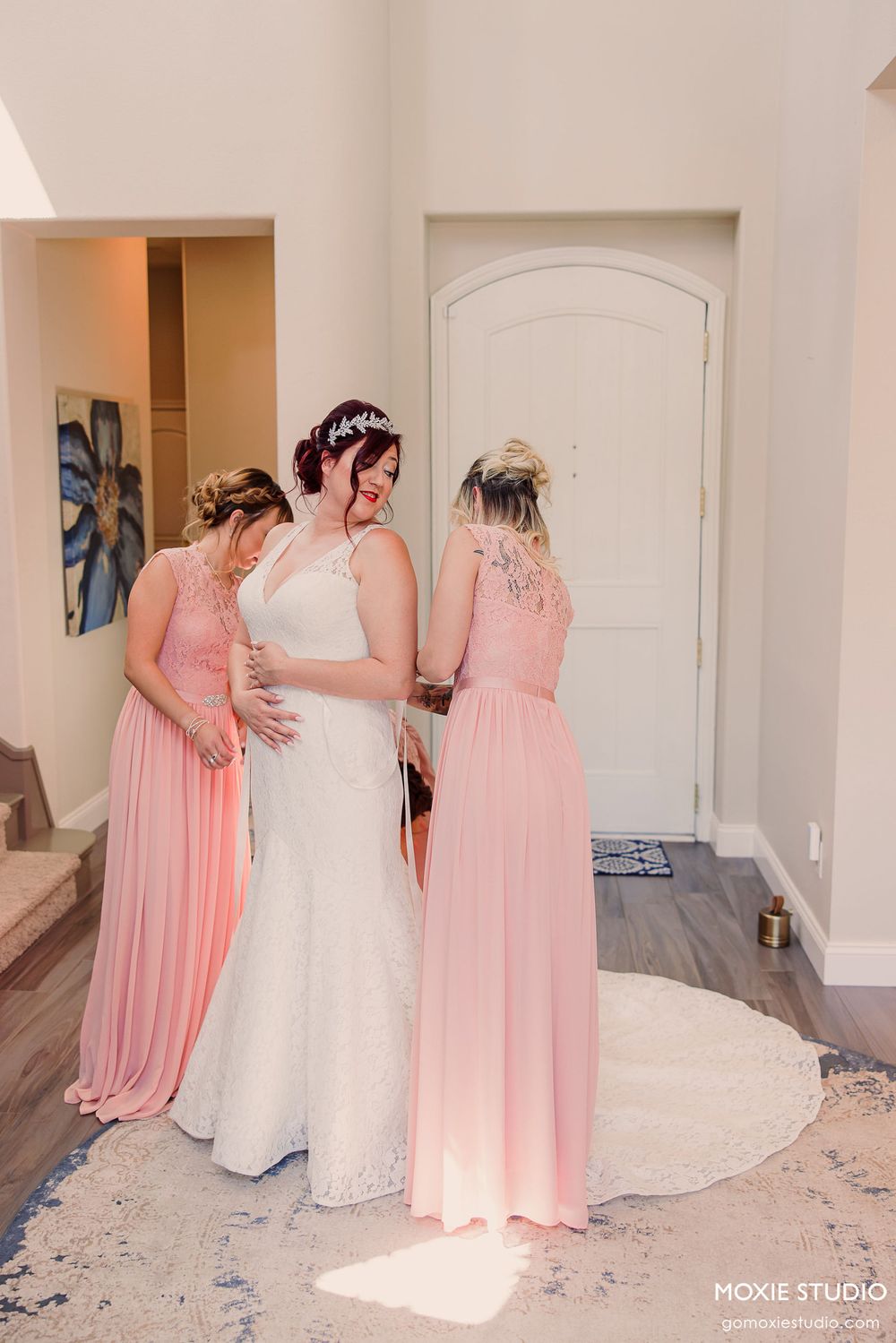 Bridesmaids in coral pink dresses help the bride get ready in an elegant white room before the wedding ceremony.