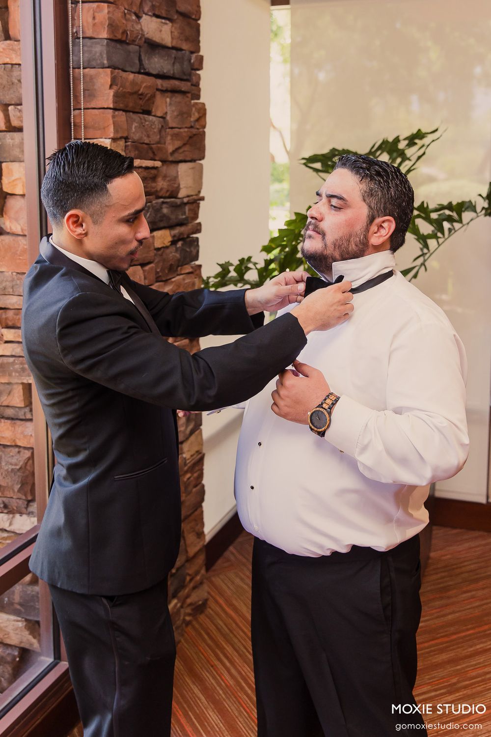 Three sequential photos show two men in formal attire adjusting a bowtie in a room with brick accent walls.