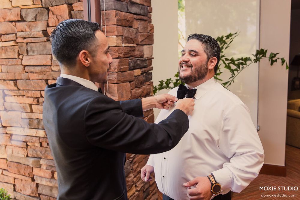 Groomsmen help adjust the groom's white tuxedo jacket in front of a rustic stone wall.