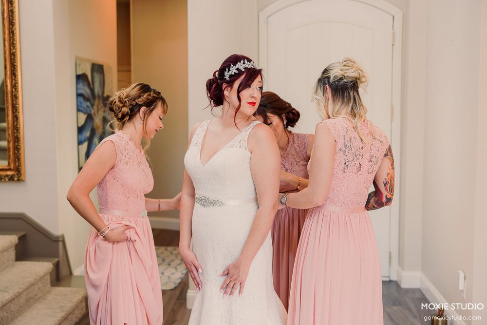 A wedding party poses in elegant pink bridesmaid dresses and a white bridal gown against a neutral interior backdrop.