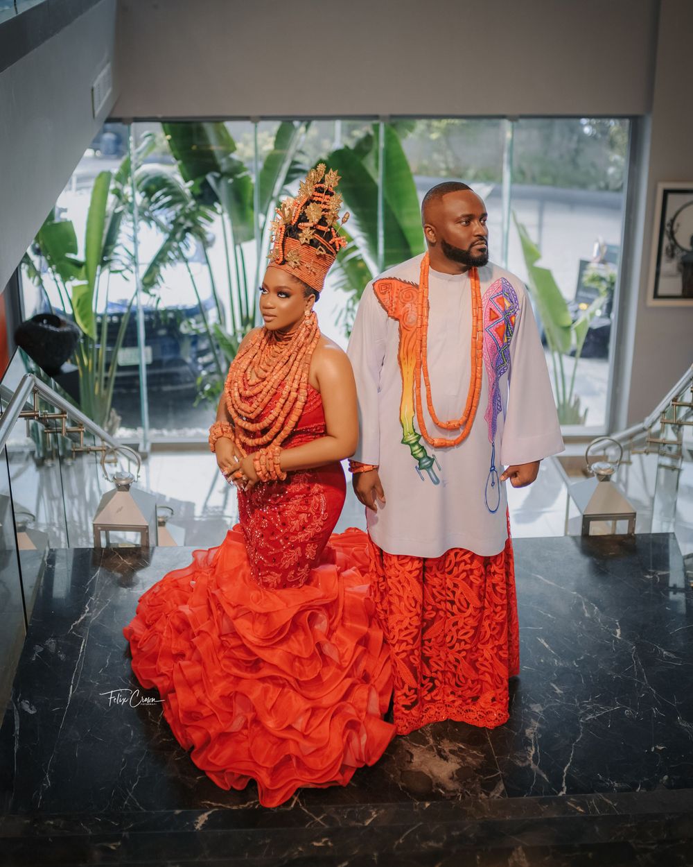 Traditional Nigerian wedding attire featuring a couple in vibrant coral red and white outfits with ornate beaded accessories.