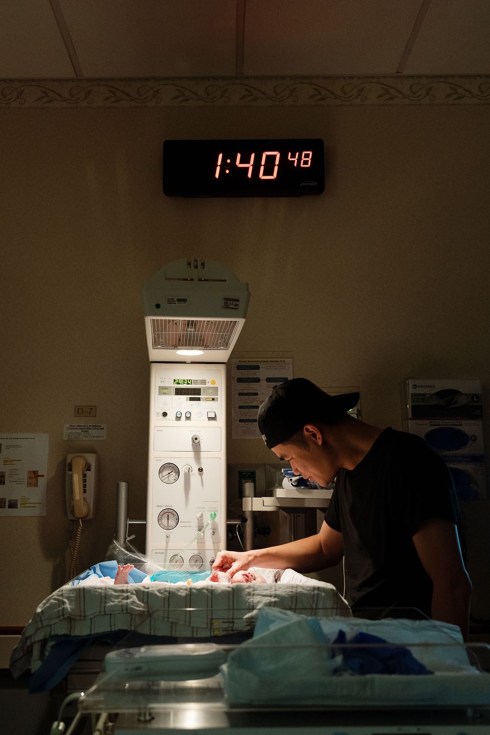 A person tends to a newborn in a hospital bassinet under a digital clock reading 1:40 in a dimly lit NICU room.
