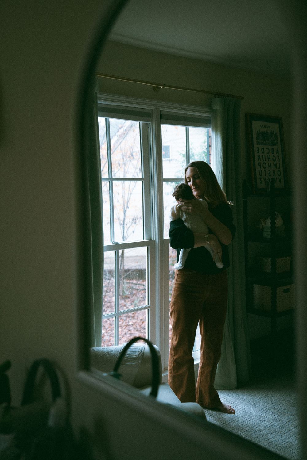 Mother holding newborn baby while standing by window, reflected in mirror.