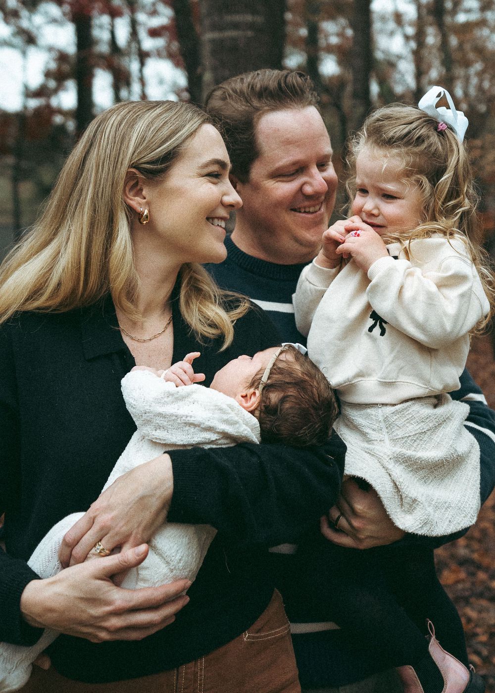Smiling family with newborn baby and toddler girl posing outdoors among autumn trees.