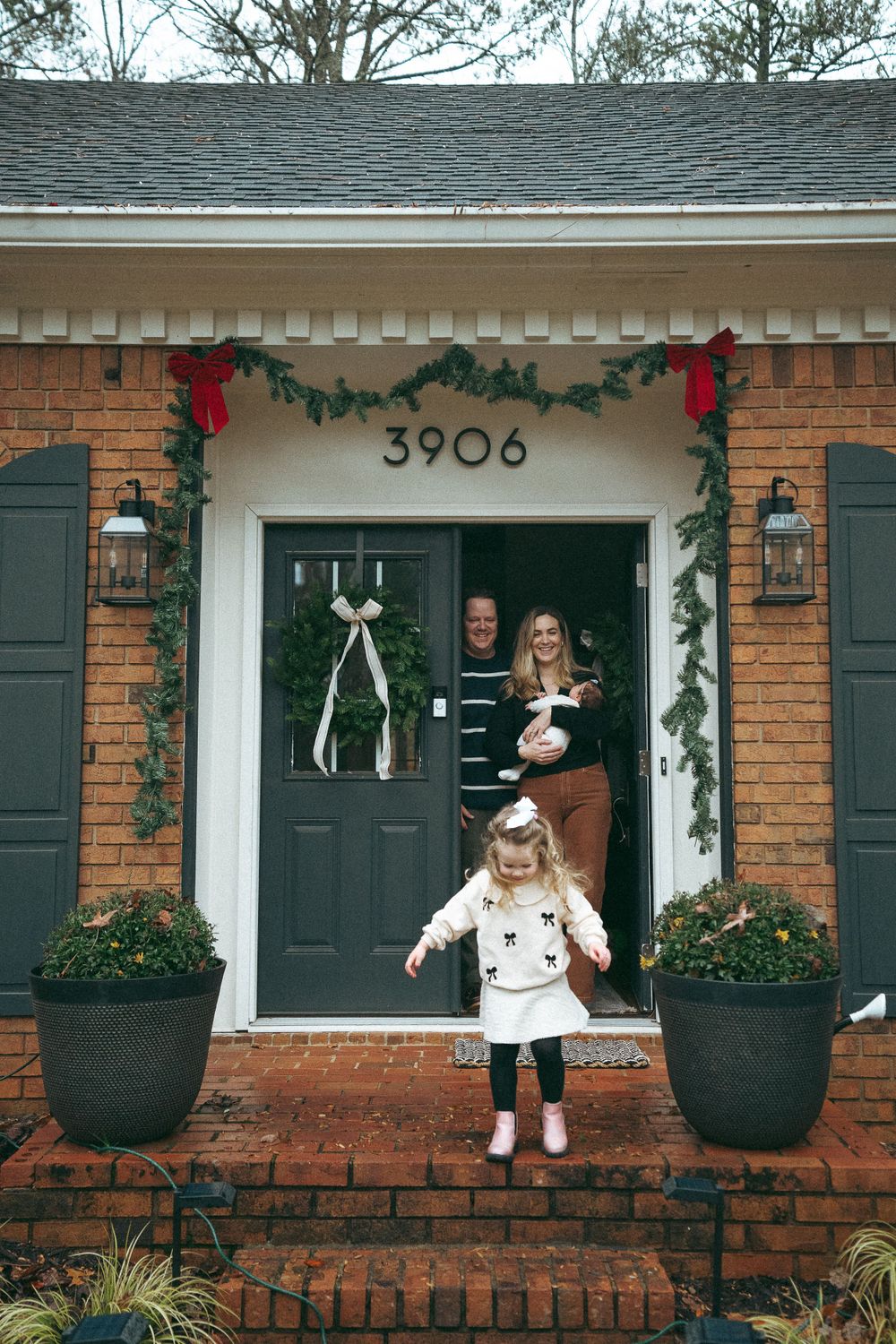 Family of four posing at Christmas-decorated brick home entrance numbered 3906.
