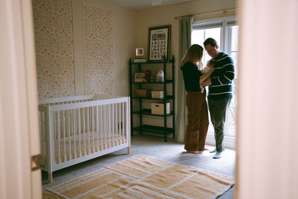 Parents holding newborn in softly lit nursery with white crib and floral wallpaper.