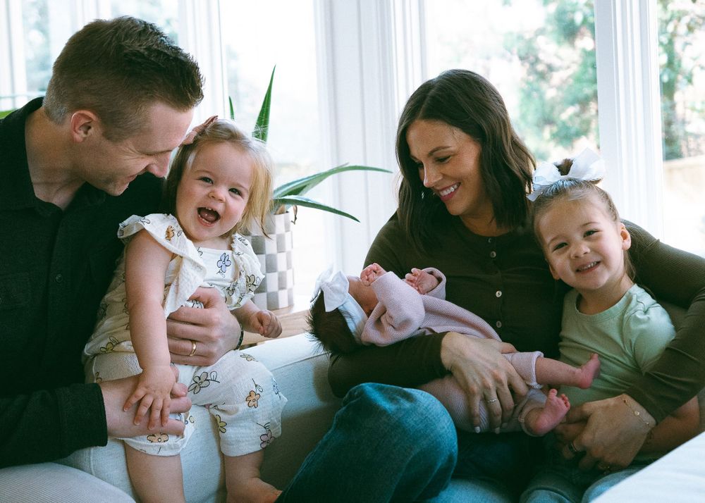 Happy family of four with two young daughters and newborn baby sitting together on white couch.