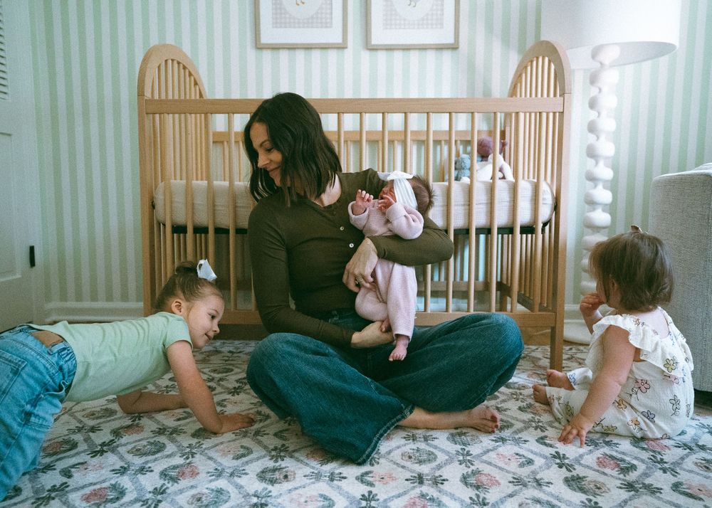 Mother sitting cross-legged on nursery floor holding newborn while two toddler girls play around her.