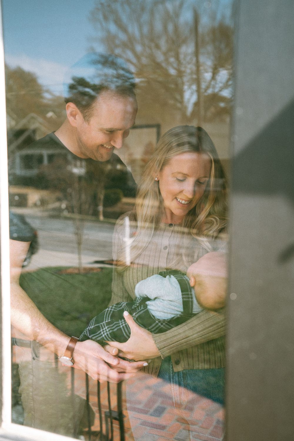 Couple smiling at newborn baby photographed through reflective window.