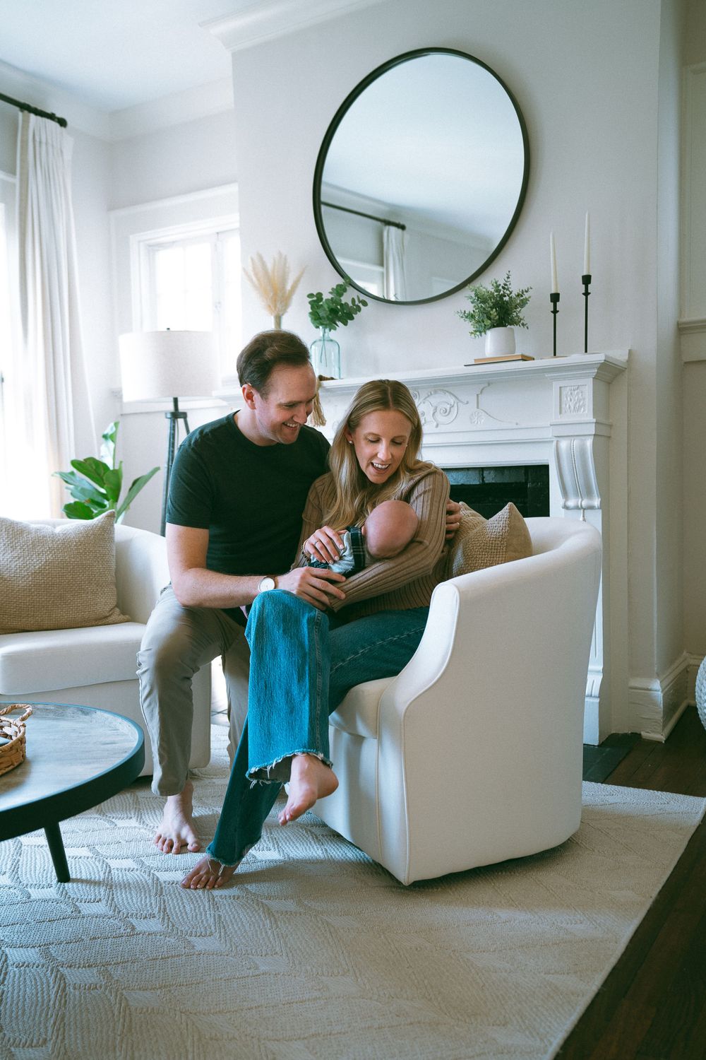Couple sitting together in a bright living room, lovingly looking at their newborn baby in a white armchair.