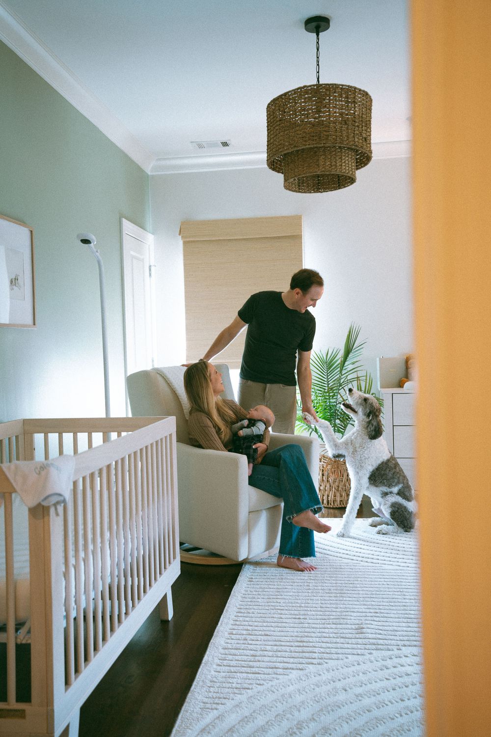 Father standing over mother sitting in nursery glider chair holding newborn baby beside white crib.