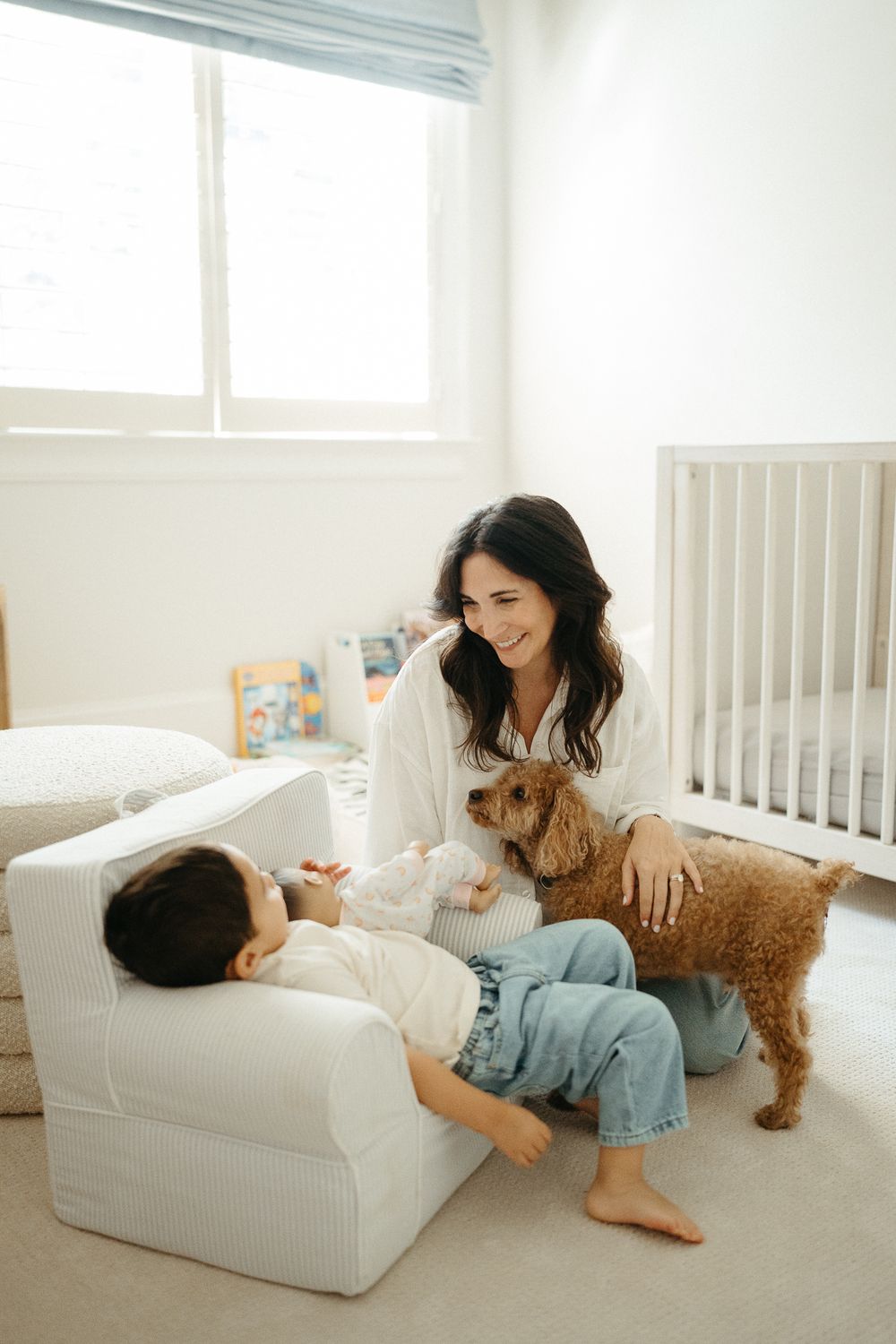 A smiling mother sits on the floor with her toddler and golden doodle dog in a bright, white nursery room.