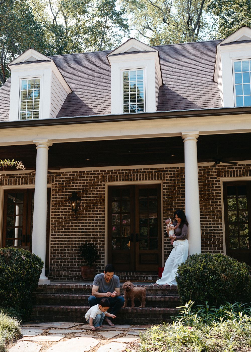 Family with baby and dog relaxing on the front porch of a charming brick colonial home surrounded by lush trees.