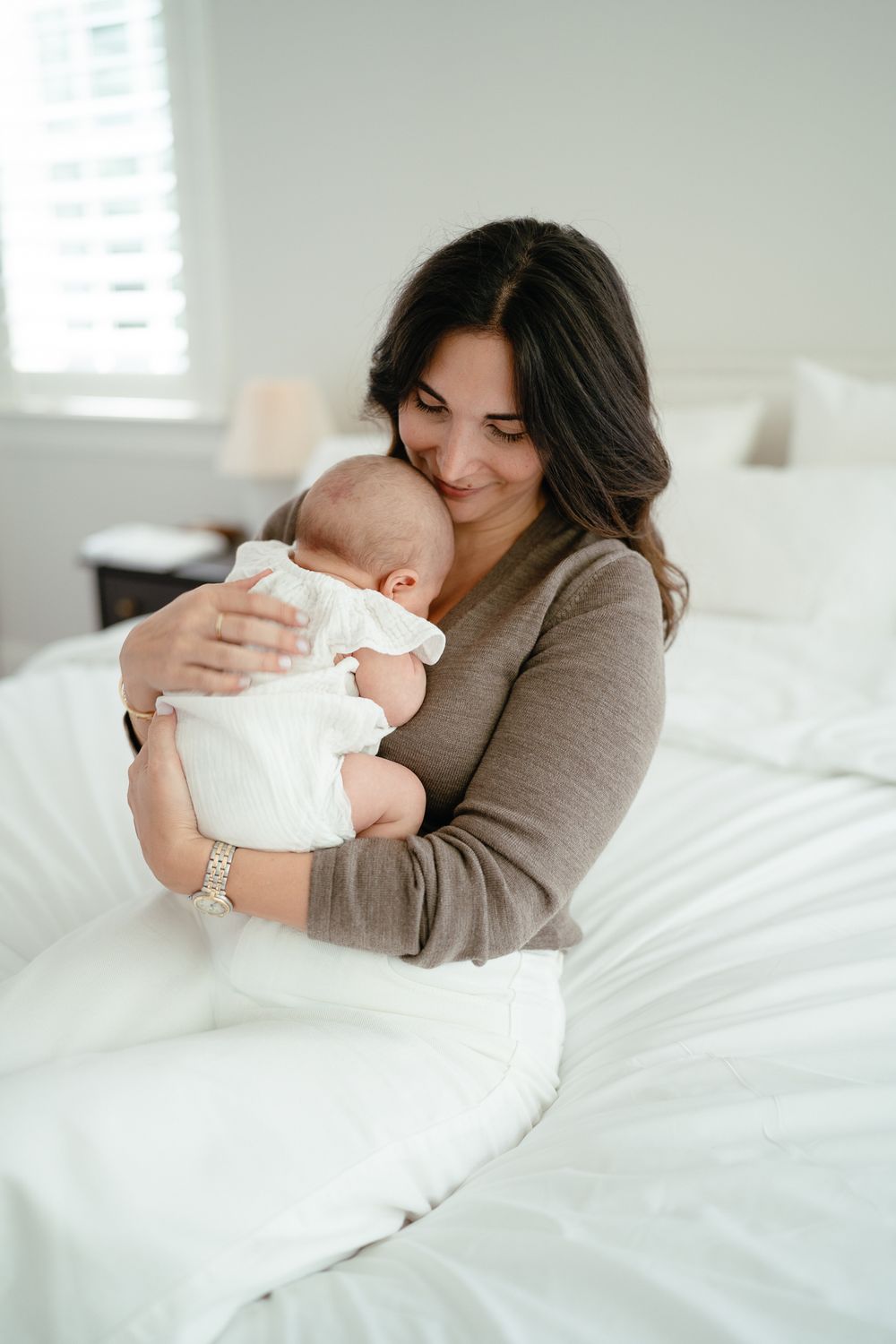 A smiling mother in a gray sweater tenderly holds her newborn baby while sitting on a white bed.