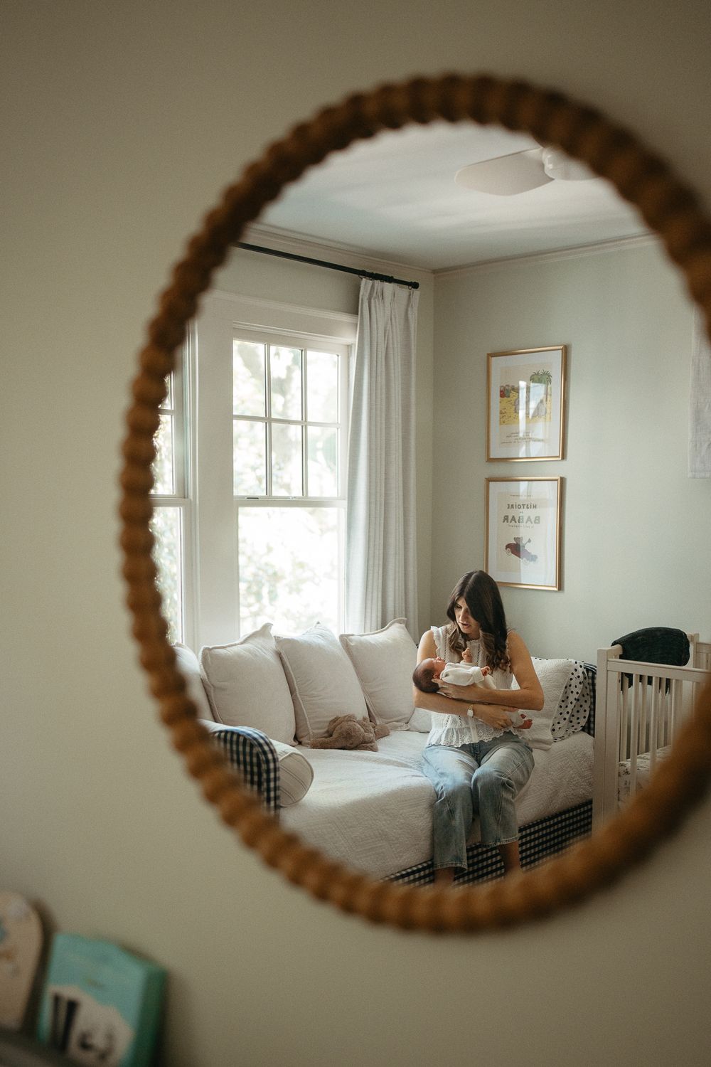 A mother sits on a cozy sofa holding her newborn baby, reflected in a round rattan mirror in a serene nursery.