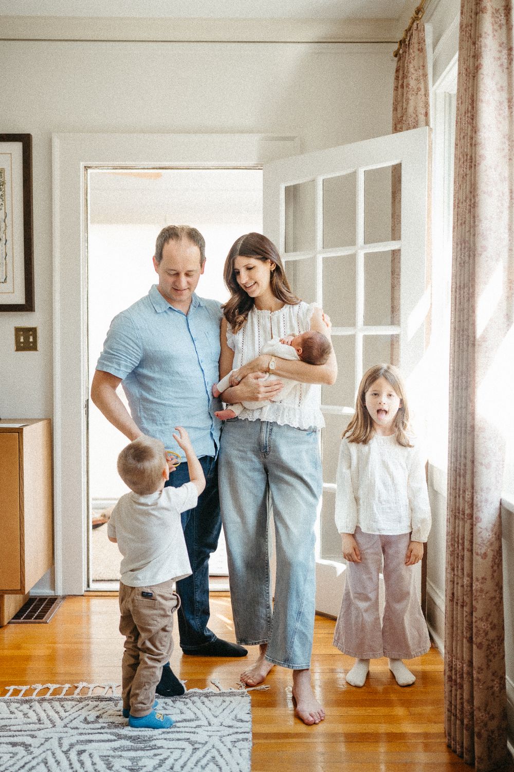 A happy family of four stands in a bright doorway, with parents holding a newborn and a toddler reaching up.