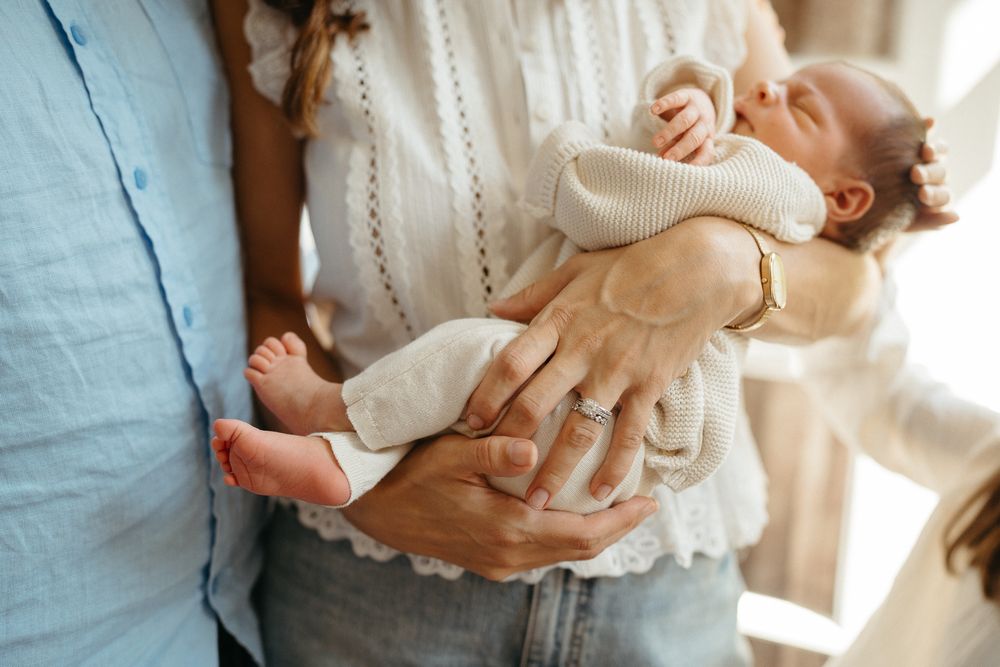Parents cradling a sleeping newborn baby in their arms, with a toddler reaching up nearby.
