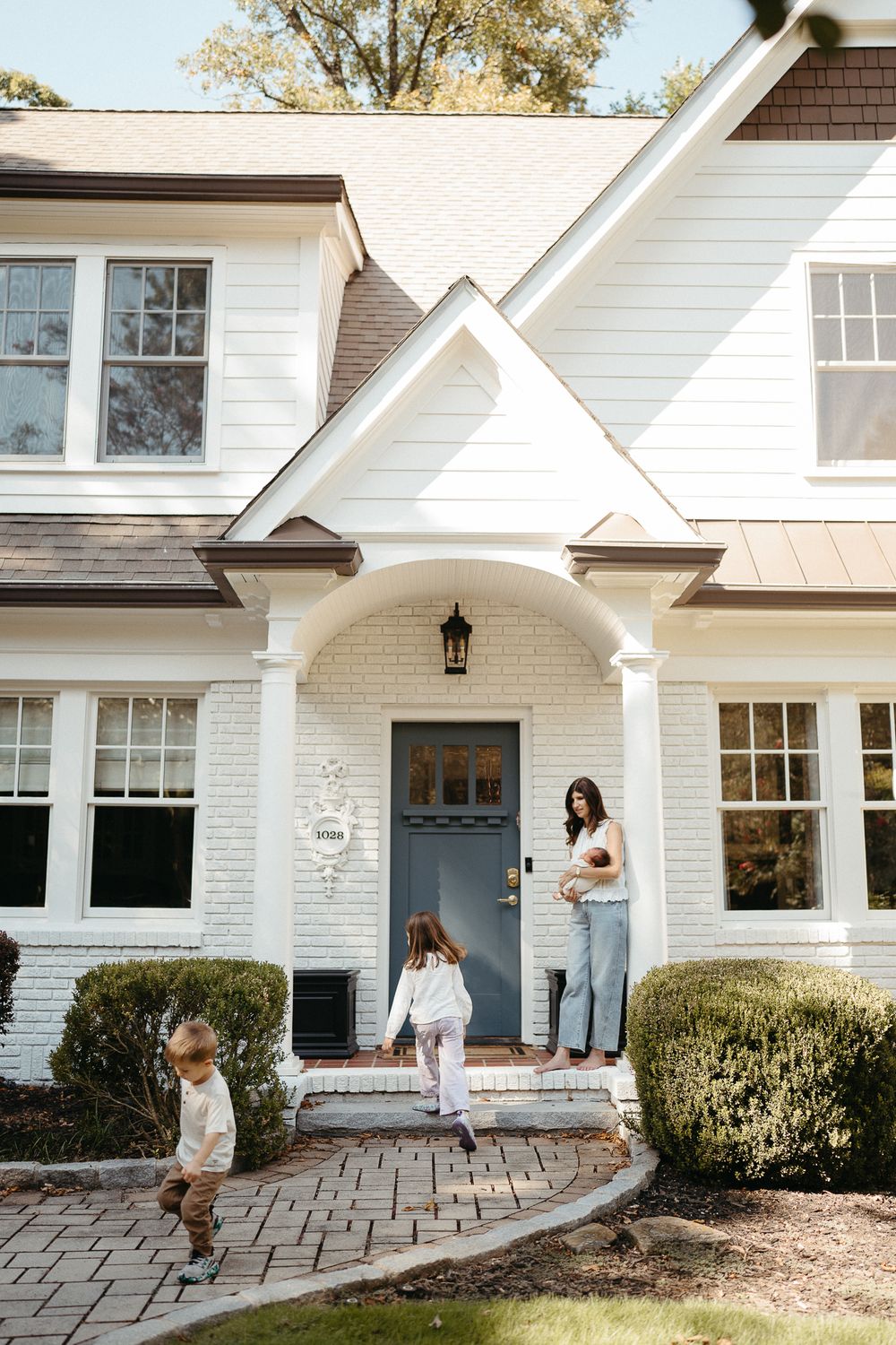 A mother holds a newborn while two young children play outside a charming white brick home with a blue front door.