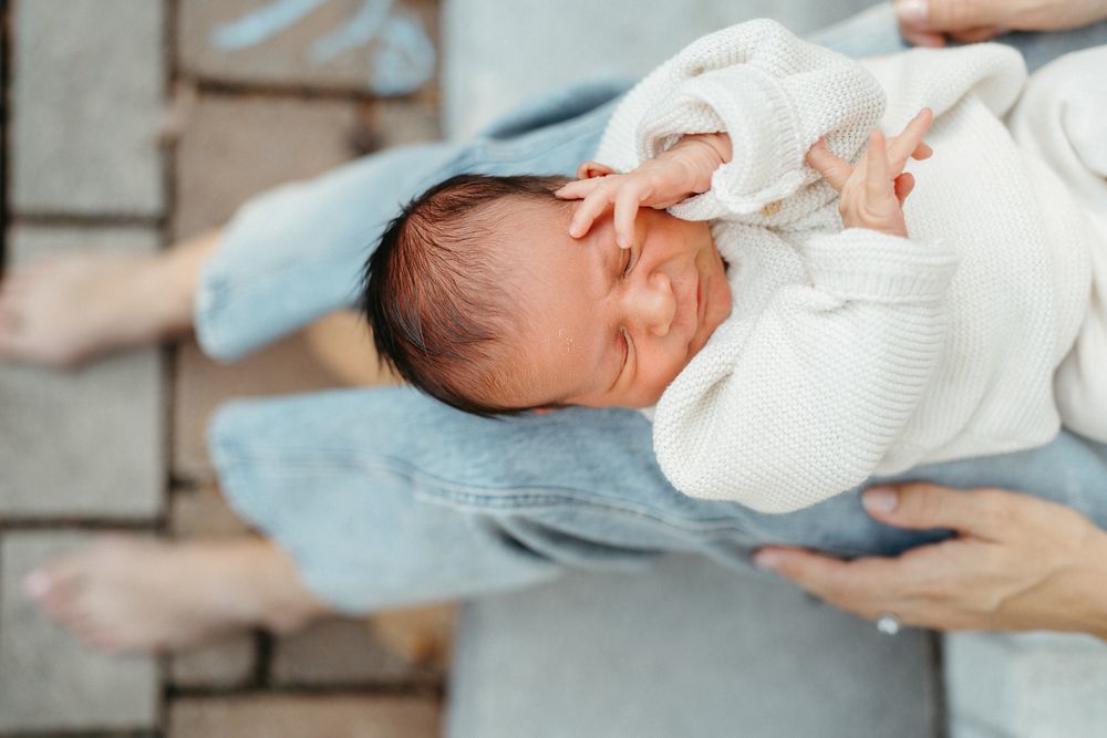 A newborn baby in a white knit outfit is cradled in a parent's arms on a brick surface.
