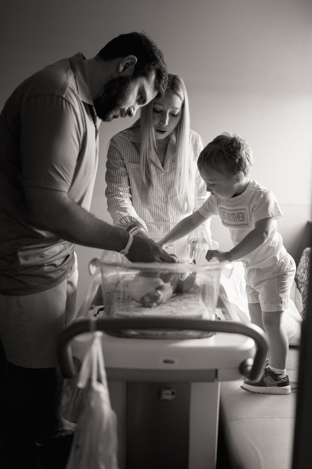Black and white photo of parents and toddler meeting their newborn baby in hospital.
