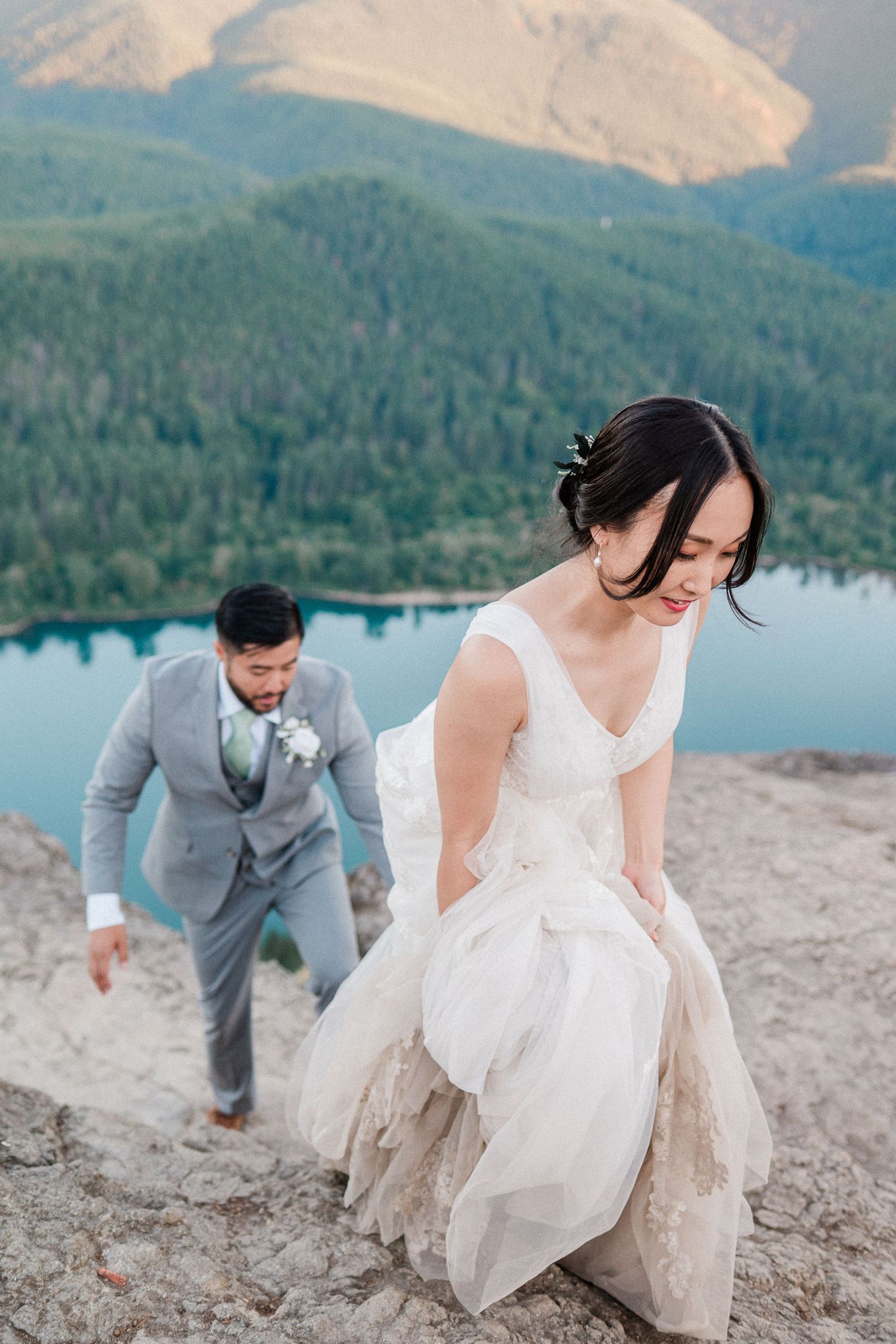 Couple hiking up mountain with bright blue lake behind them in wedding attire. photo by Pine + Vow