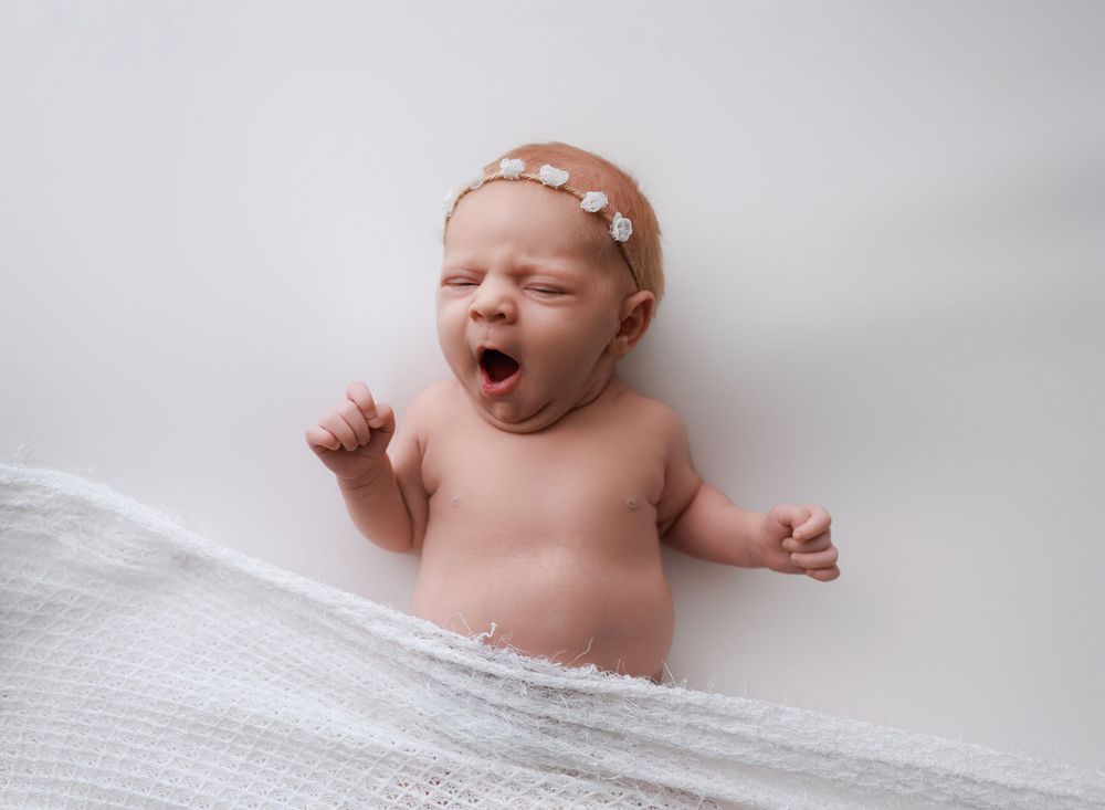A newborn baby yawning while lying on soft white textured fabric in a studio setting.