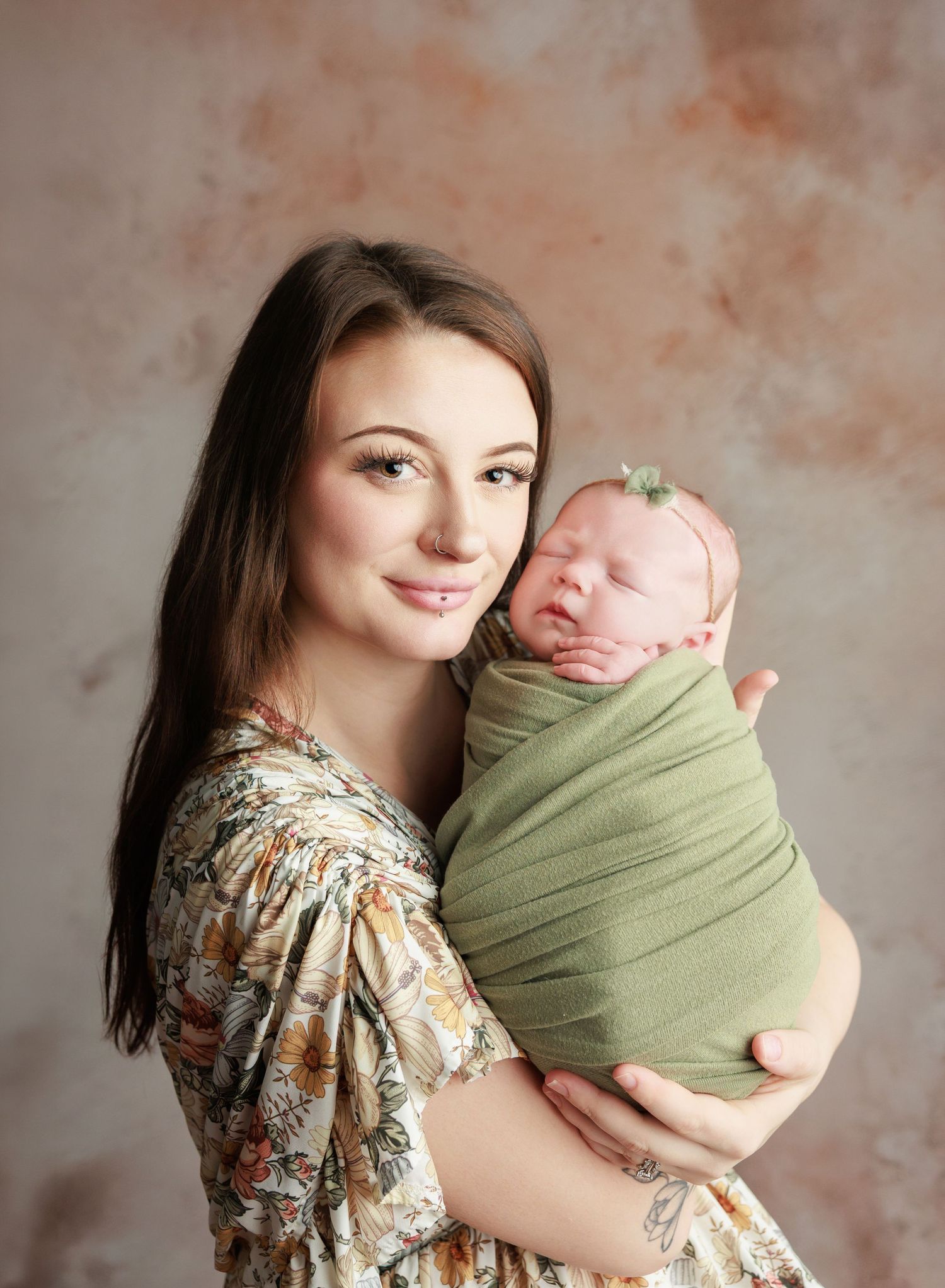 A young mother in a floral dress holds her swaddled newborn wrapped in green fabric for a studio portrait.
