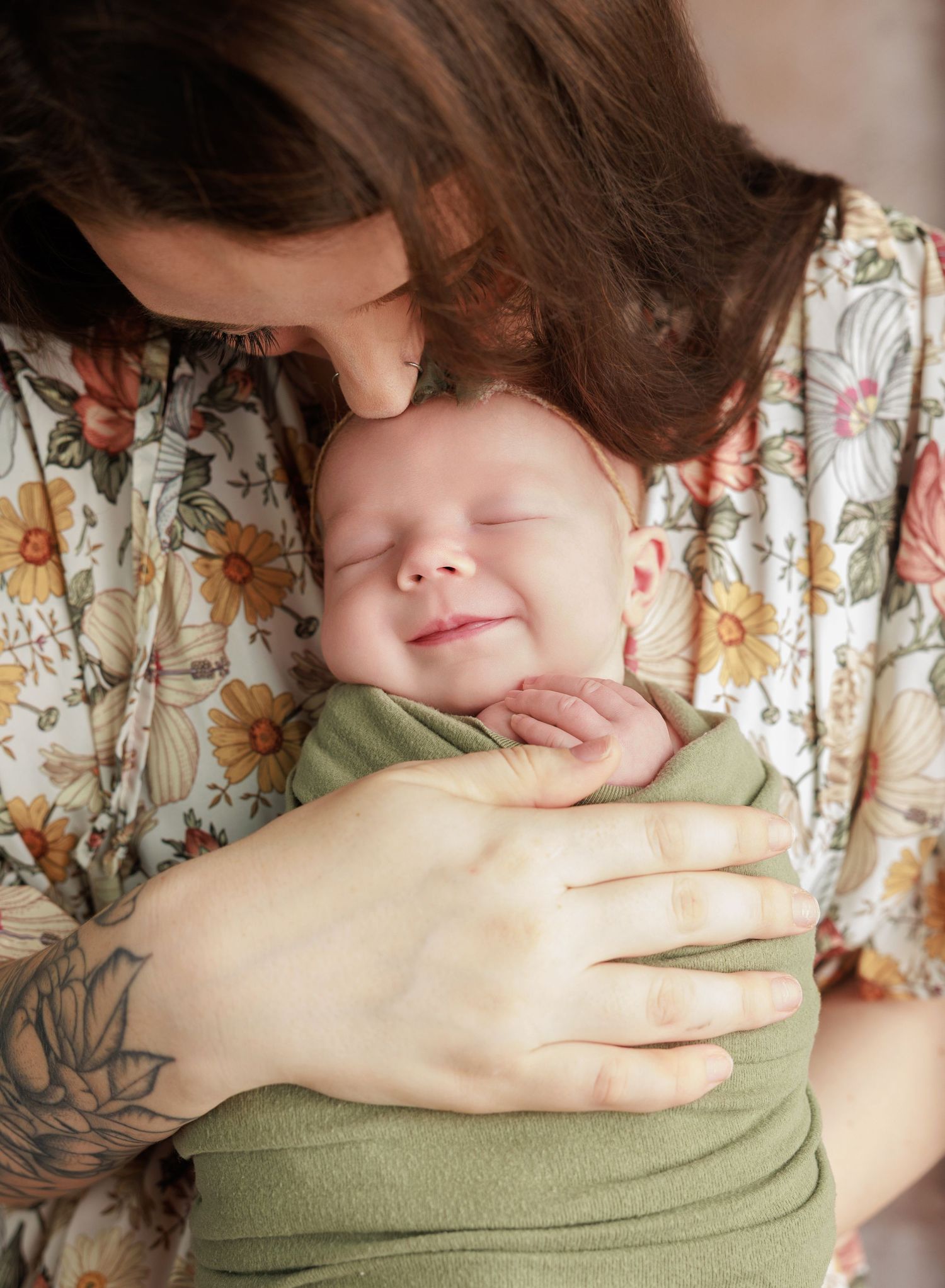 A mother with a floral dress and tattoo tenderly kisses her smiling newborn baby wrapped in a green swaddle.
