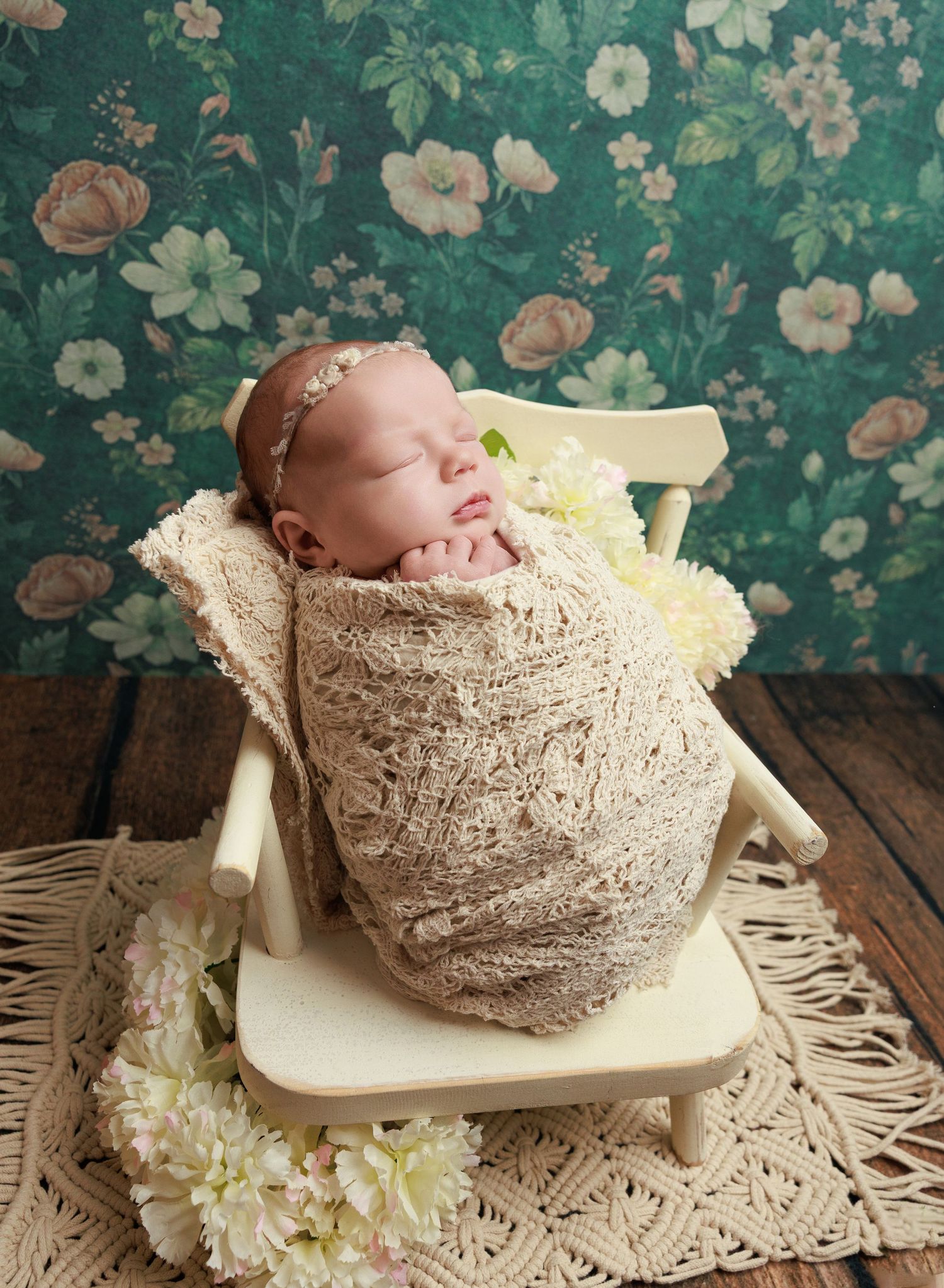 Sleeping newborn baby swaddled in beige lace wrap, posed in tiny white chair with flowers, against floral teal backdrop.