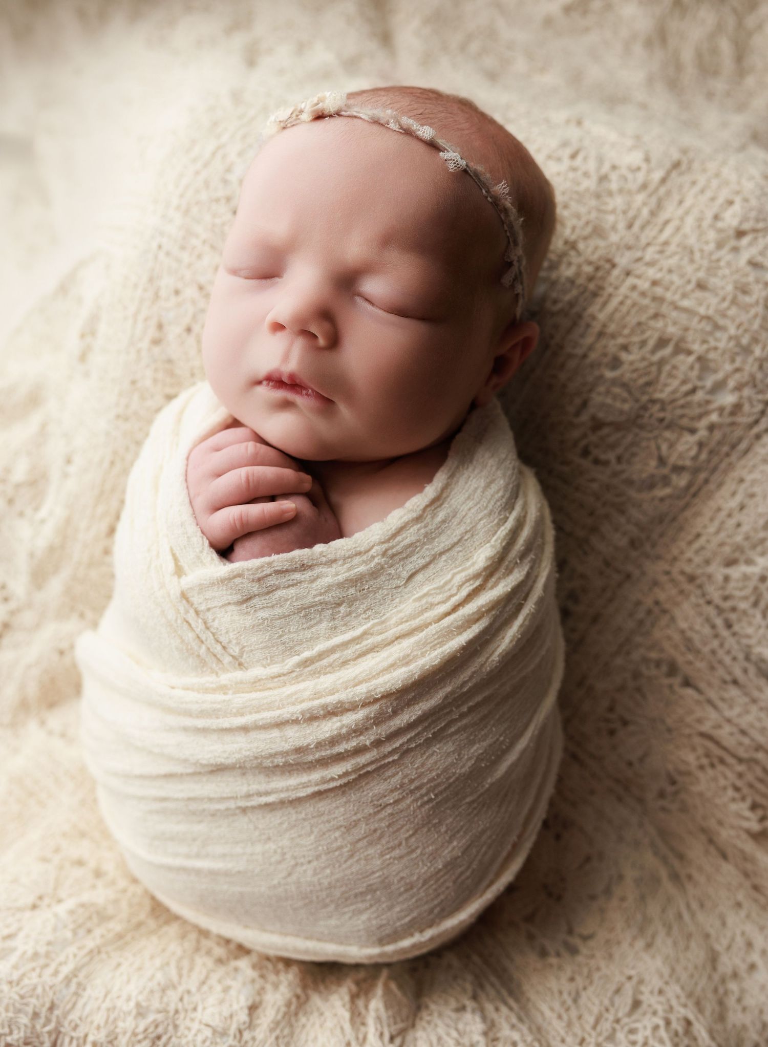 Sleeping newborn baby swaddled in cream wrap with delicate headband on soft textured blanket.
