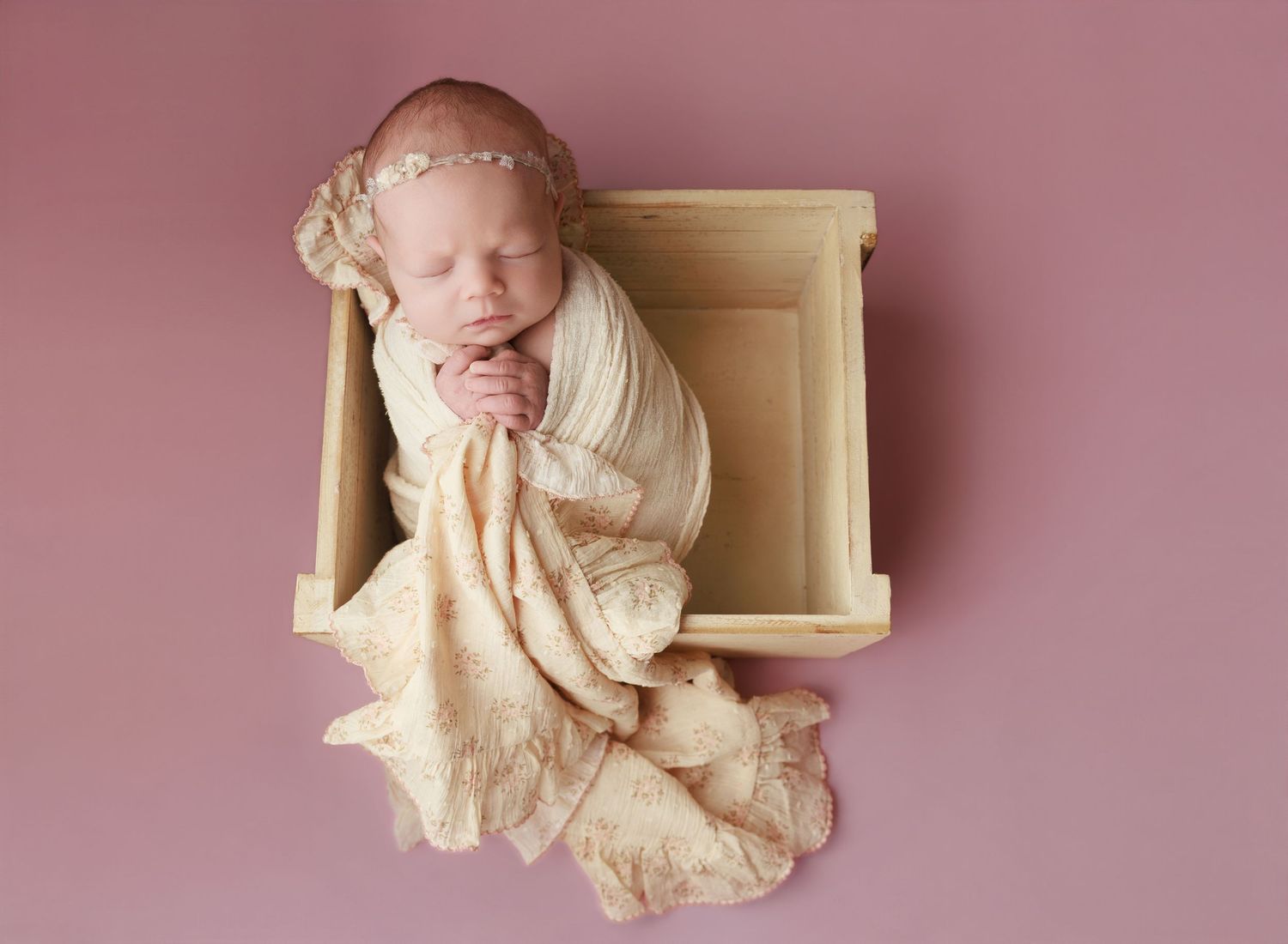 Newborn baby wrapped in cream swaddle sleeping peacefully in a wooden crate on a pink background.