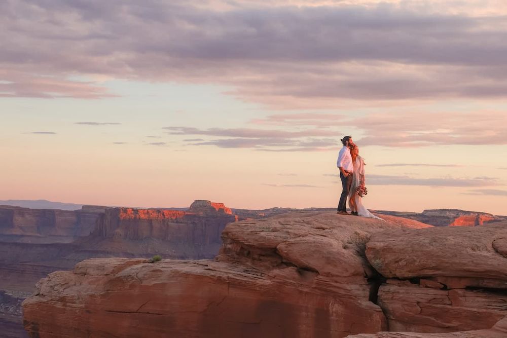 Couple embracing on a red rock cliff edge at sunset overlooking a vast canyon landscape.