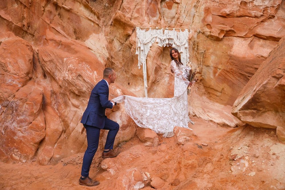 Bride in lace gown and groom in blue suit pose among red rock canyon formations during desert wedding elopement.
