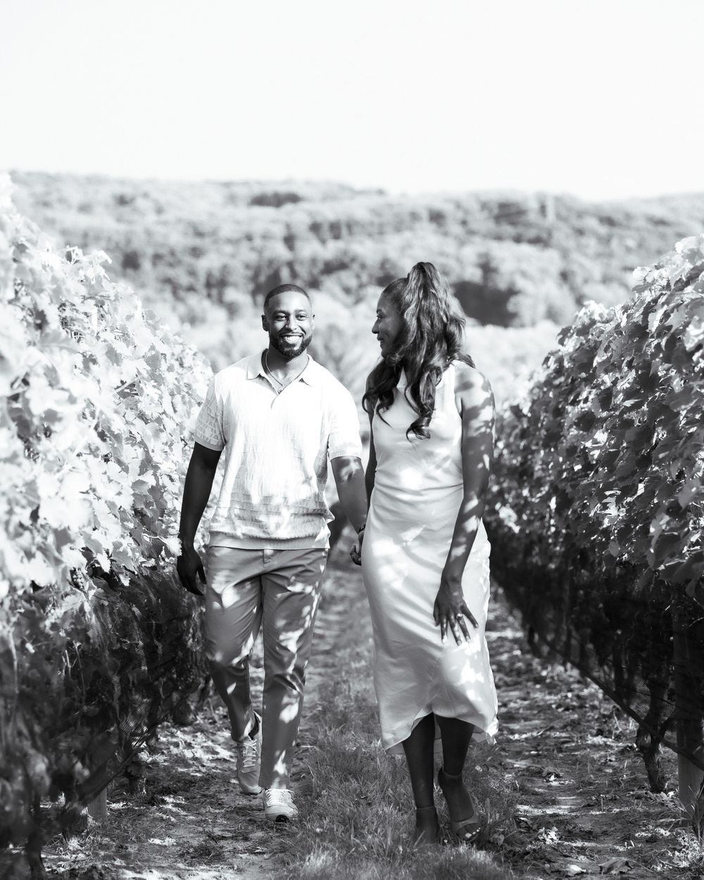 A romantic couple walks through a vineyard holding hands in black and white.