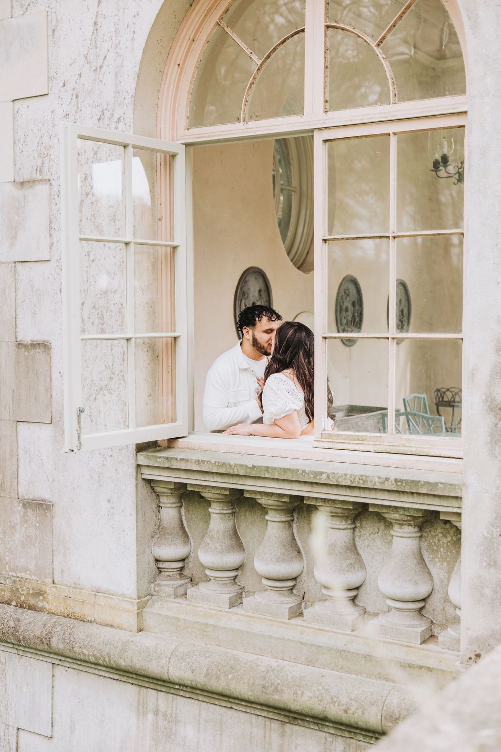 Couple sharing romantic moment through grand arched window with decorative stone balustrade.
