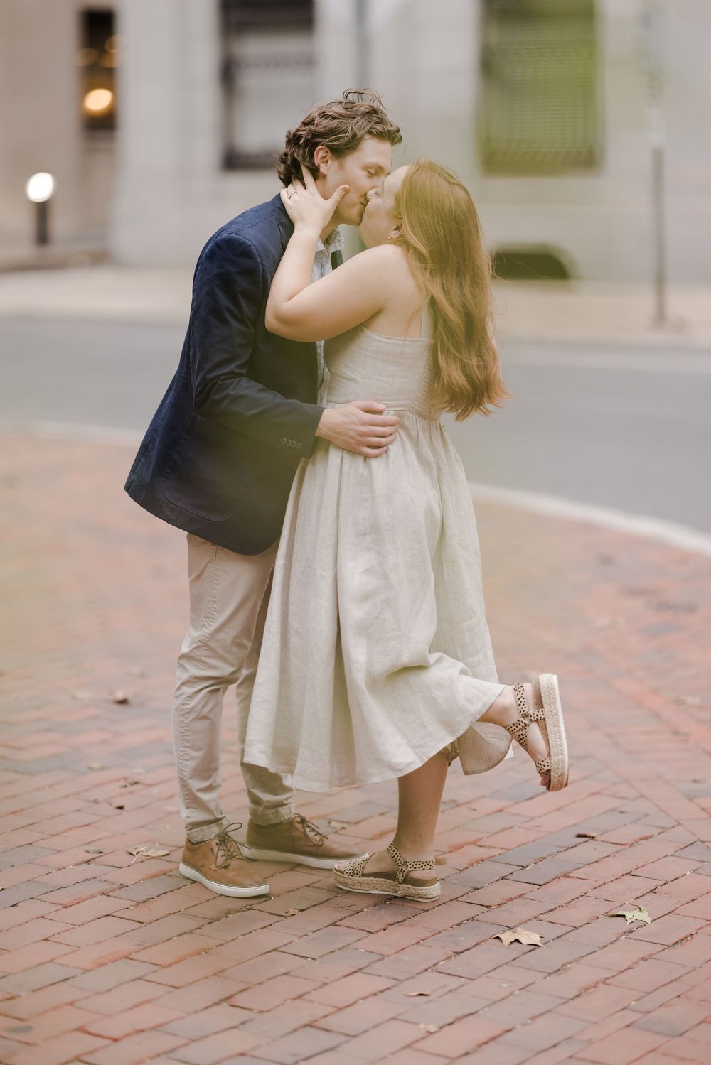 A romantic couple embraces and kisses on a brick sidewalk, with the person in a white dress lifting one foot.
