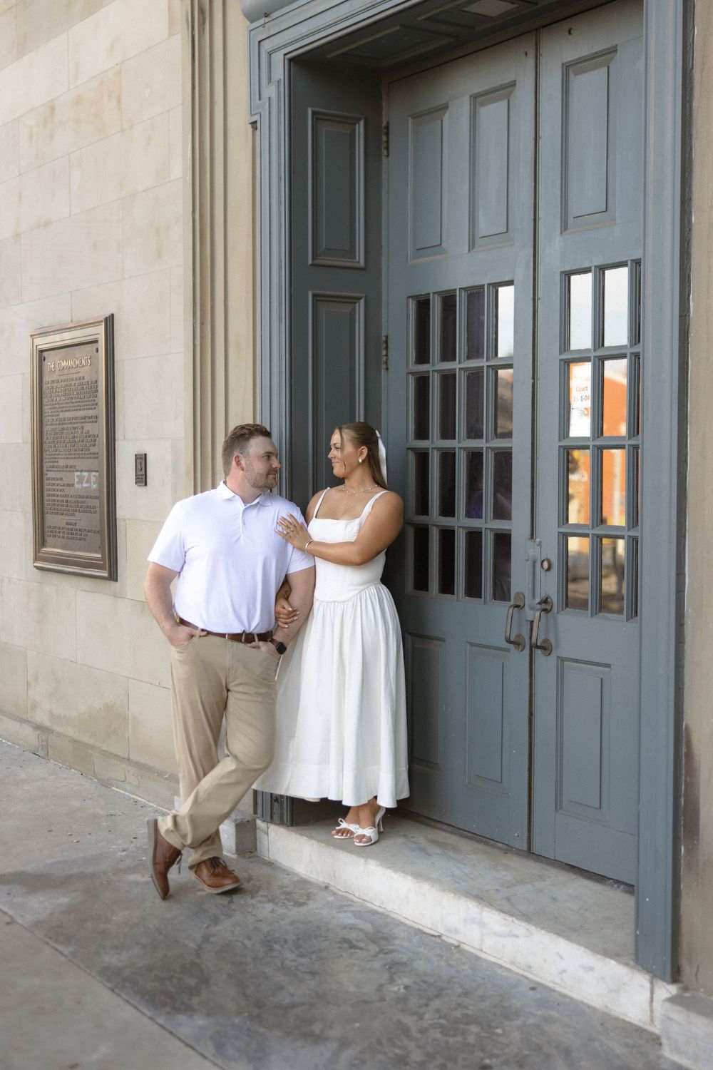 Couple posing by a blue door of a historic building, man in white polo and khakis, woman in white midi dress.