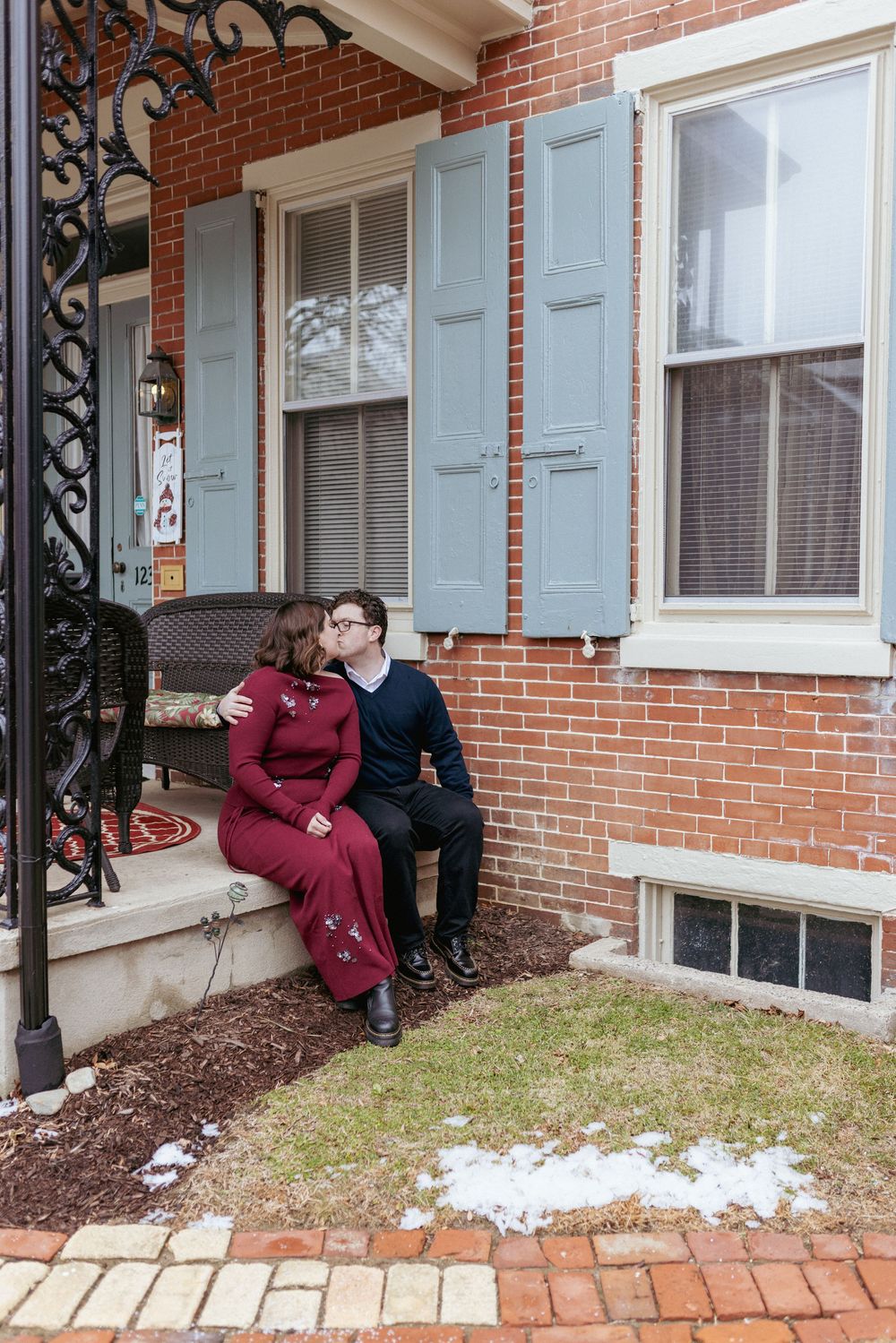 A couple sits together on brick steps outside a traditional red brick home with light blue shutters and decorative ironwork.